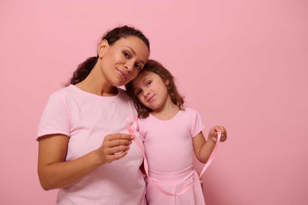 Adorable baby girl , leans on her mother shoulder,both dressed in pink color outfit, hold pink satin ribbons, cute smile looking at camera . World Breast Cancer Awareness Day. Fight Cancer. Copy space