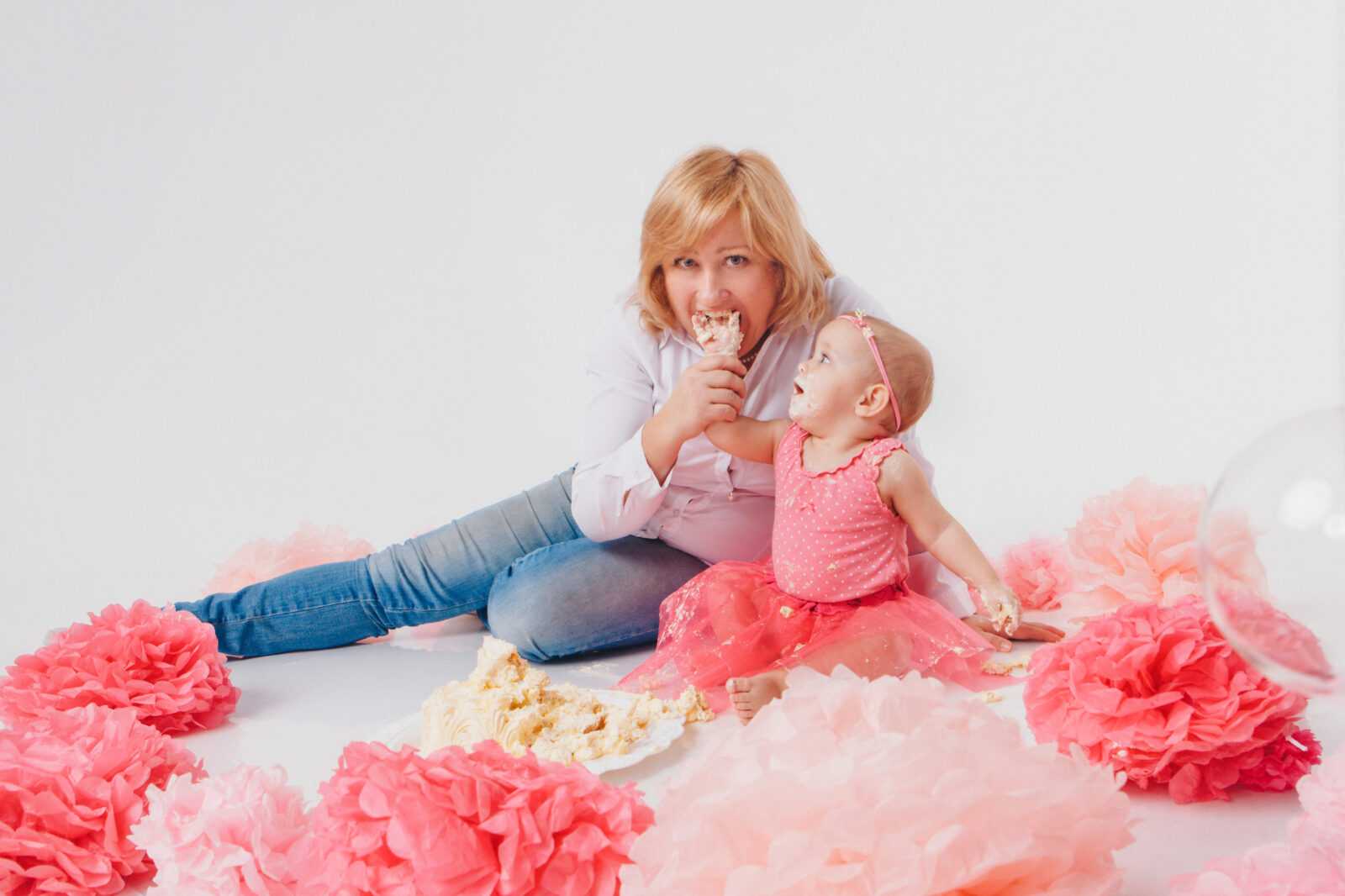 Birthday celebration: little girl eating cake with her hands on white background. The child is covered in food. Ruined the sweetness. on the floor among the decoration: numbers 1, artificial flowers and white balls